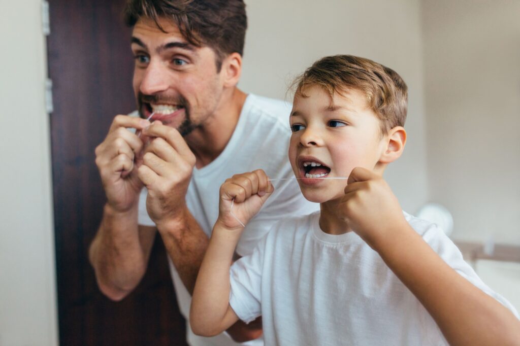 Man and boy flossing teeth together in front of bathroom mirror